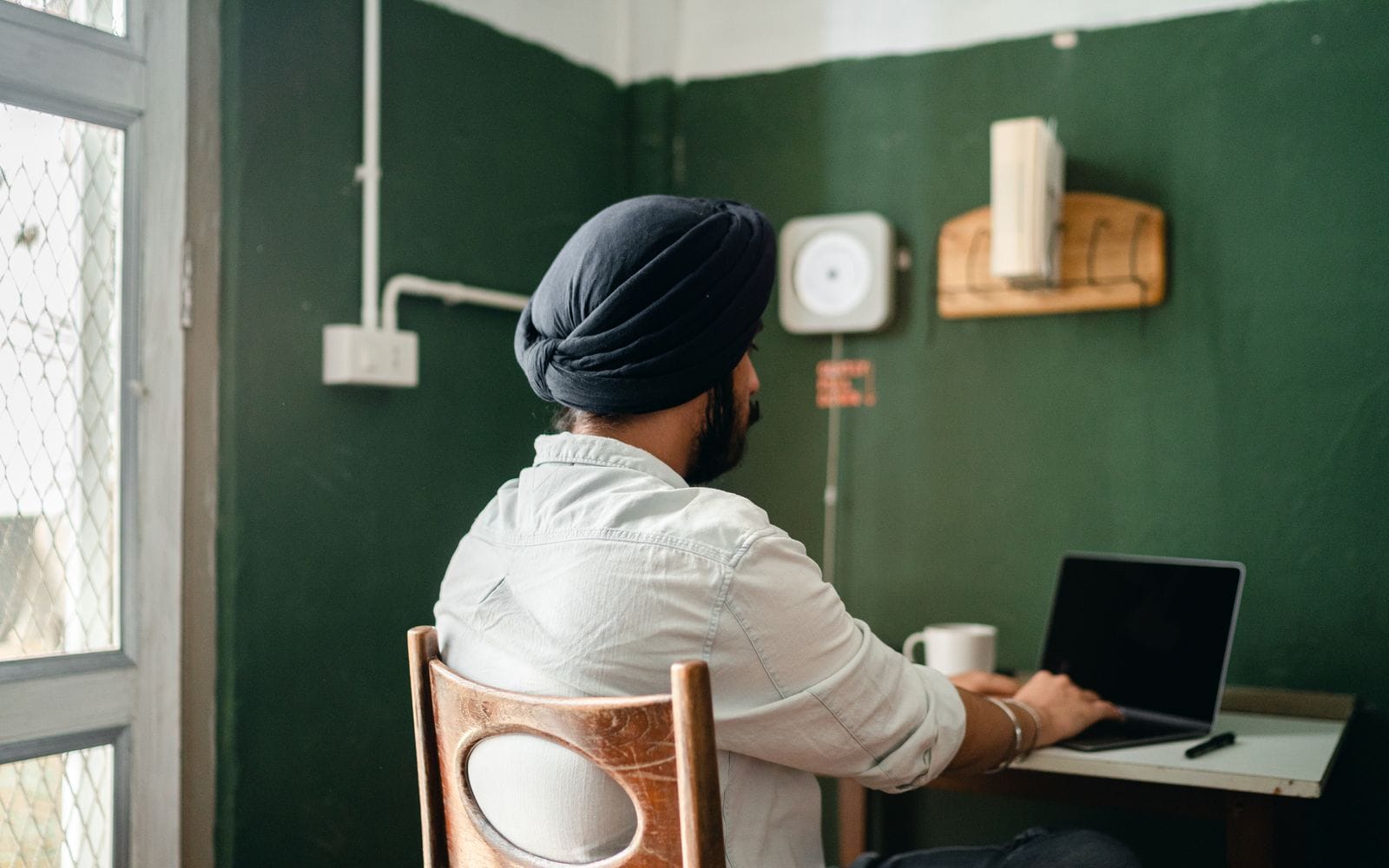 A man in a turban working on a laptop at a small desk in a green-walled room