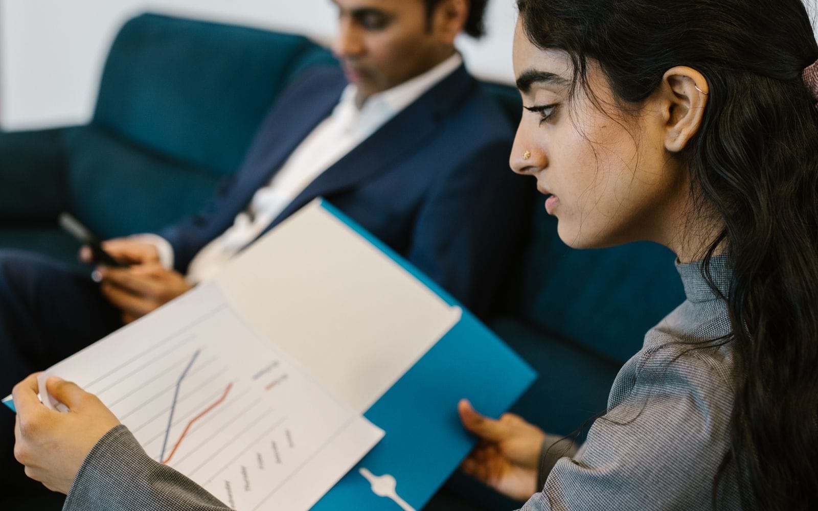 A woman reading a printed chart in an office while a colleague uses his phone