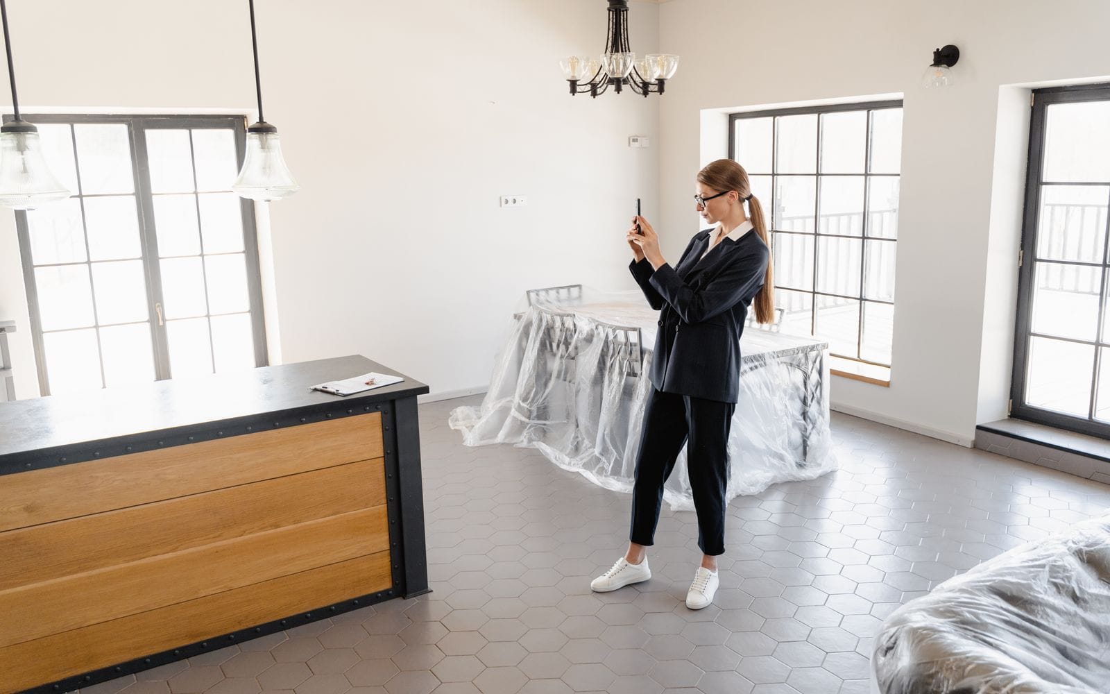 A woman in a dark suit filming an empty, bright apartment with her phone