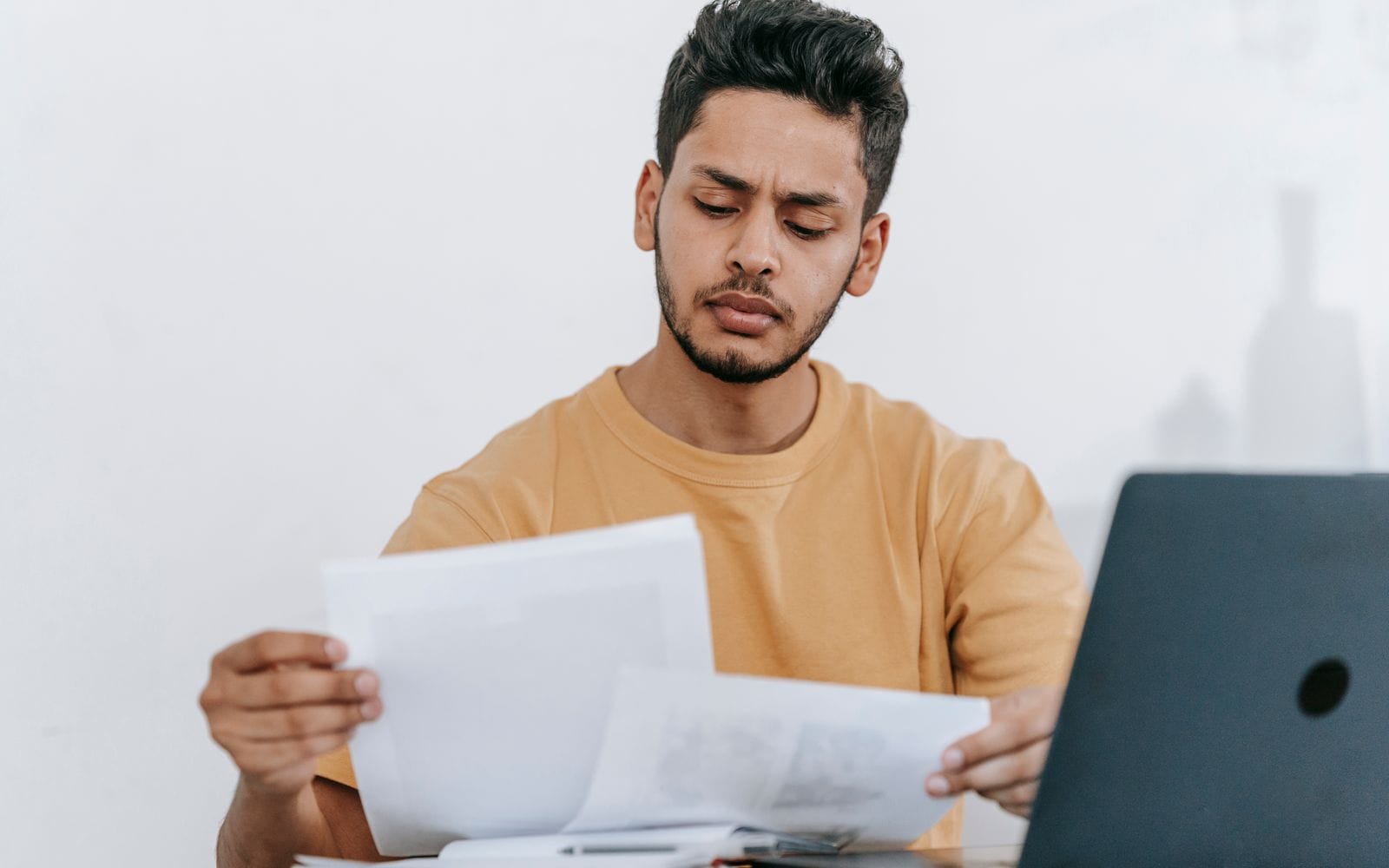 A young man in an orange T-shirt reading printed papers in front of a laptop