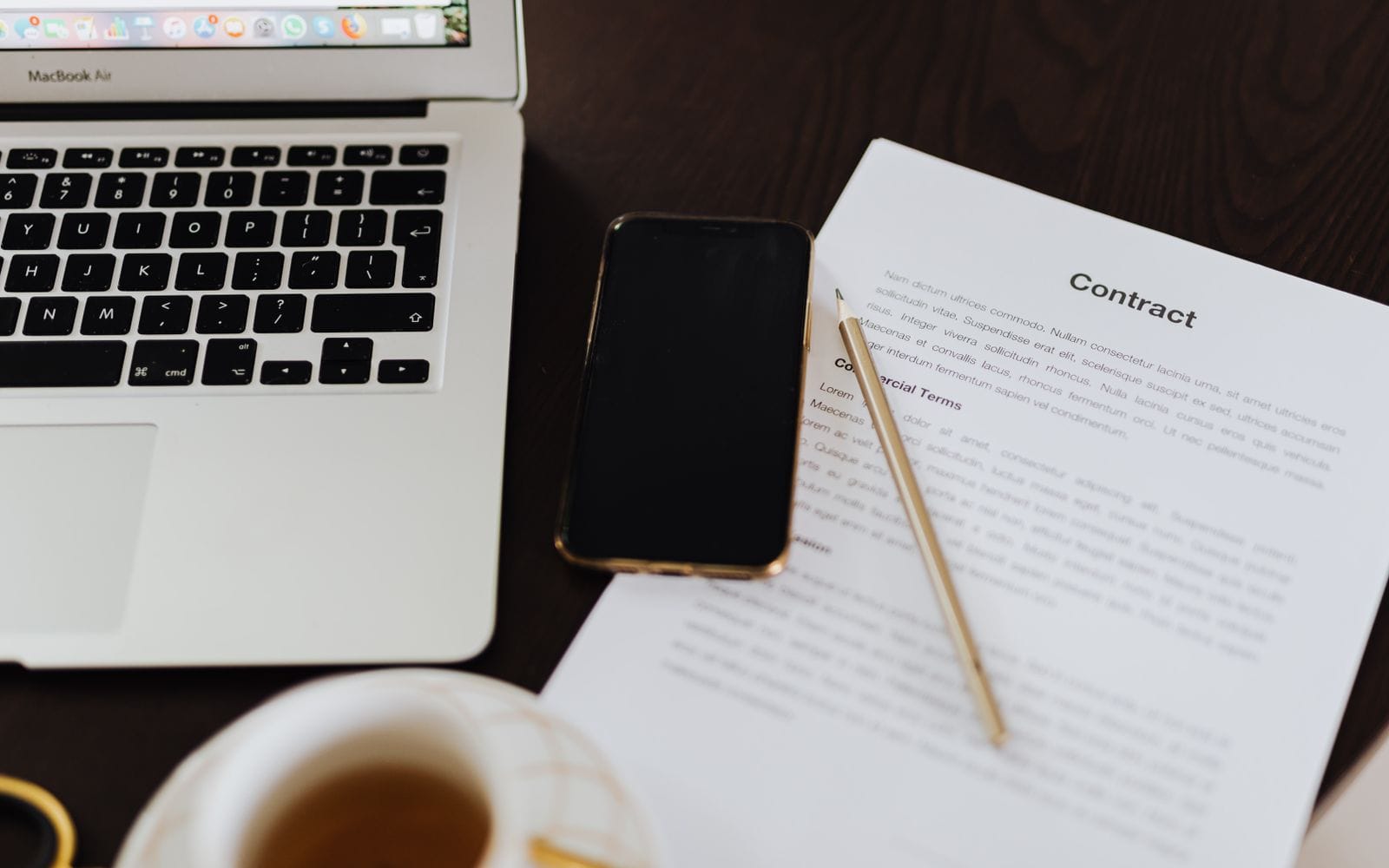 A laptop, a phone, a pen and a printed contract on a wooden desk next to a cup of tea