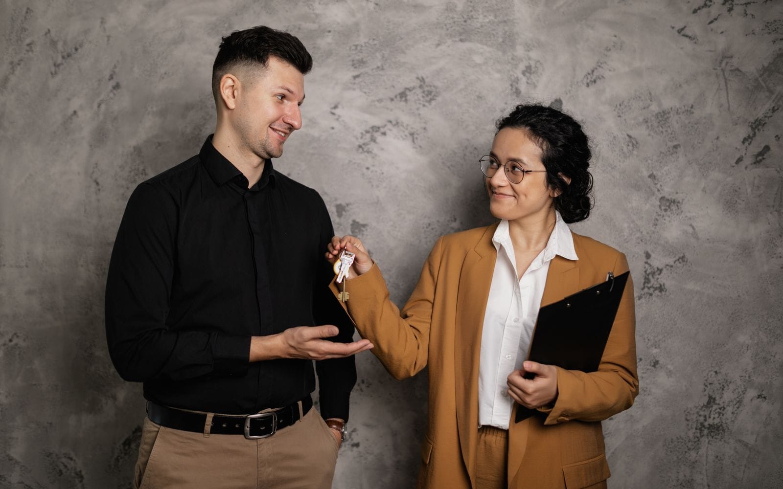 A smiling agent in a mustard blazer handing a set of keys to a client against a grey wall
