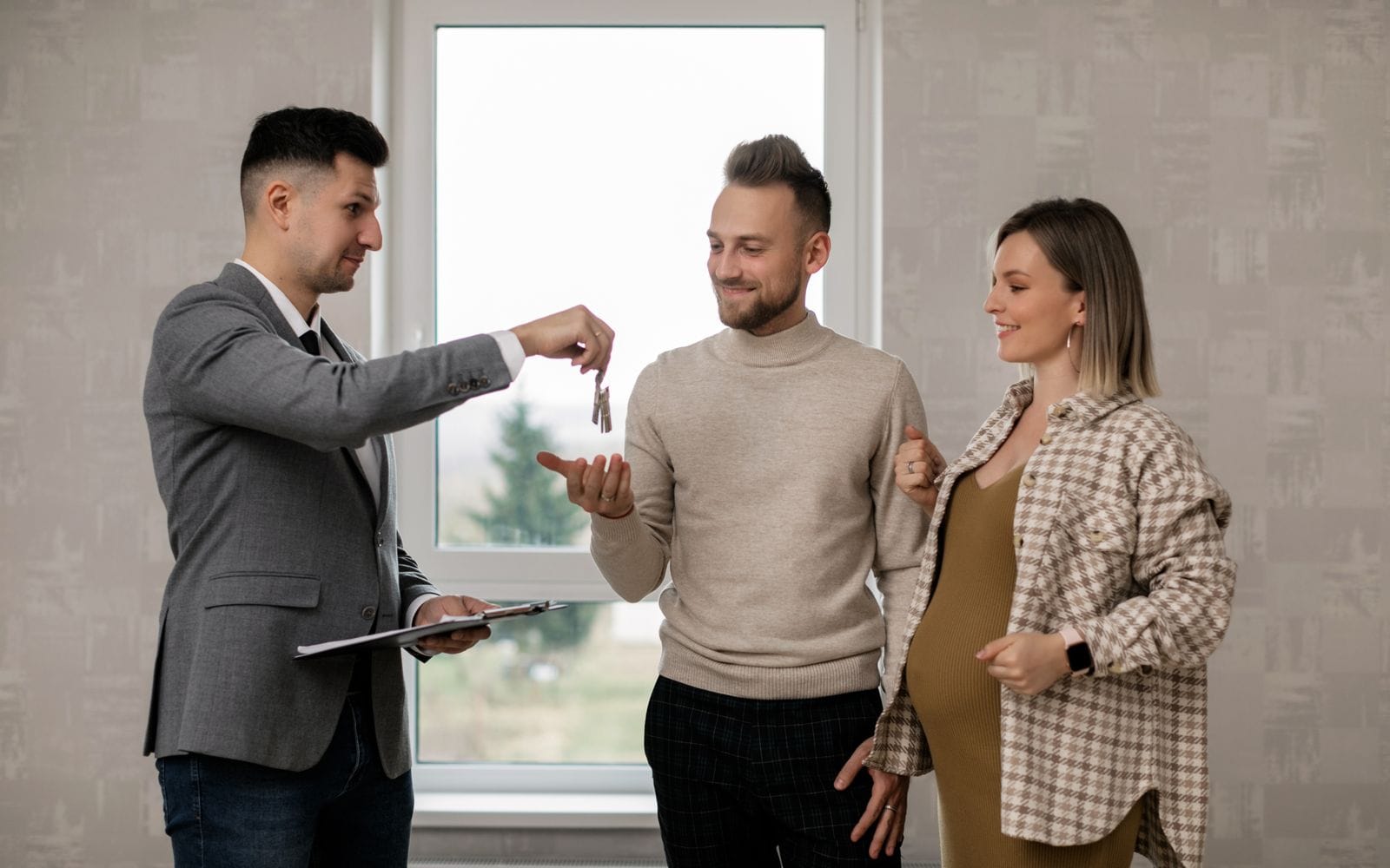 A property agent in a grey suit handing house keys to a smiling couple in a bright room