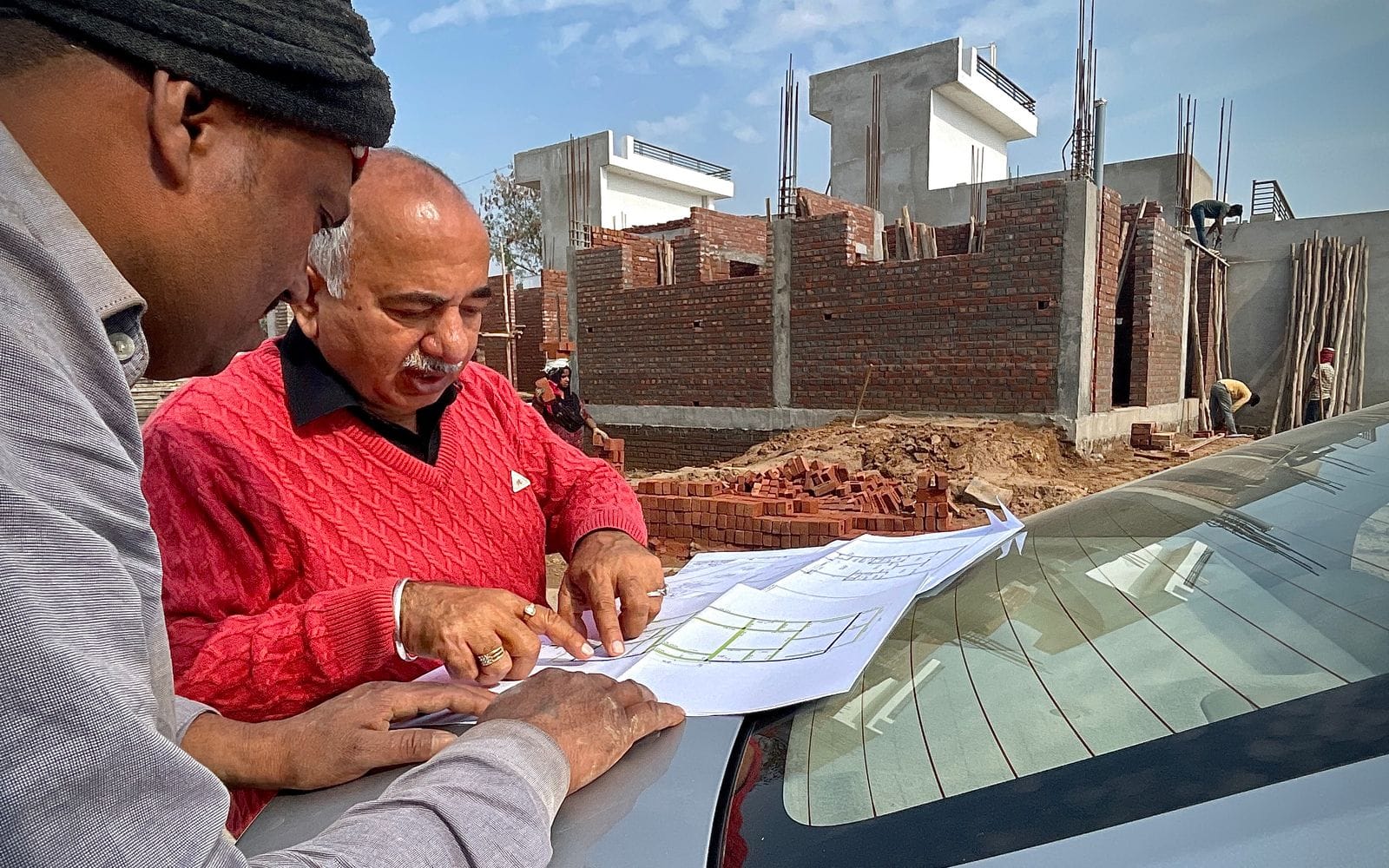 Two men reading building plans spread on a car bonnet beside a construction site