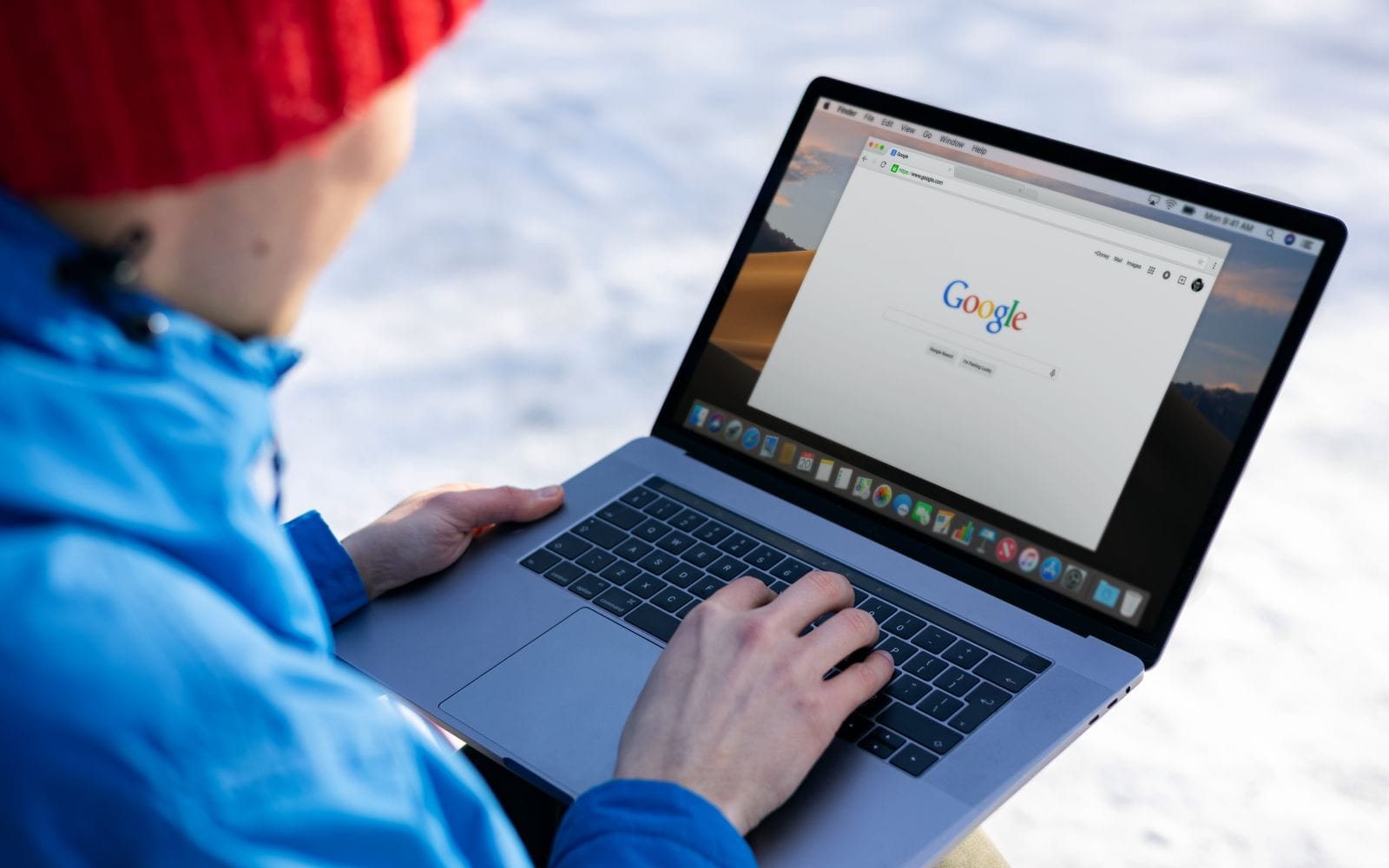 A man in a blue jacket and red beanie searching on a laptop outdoors