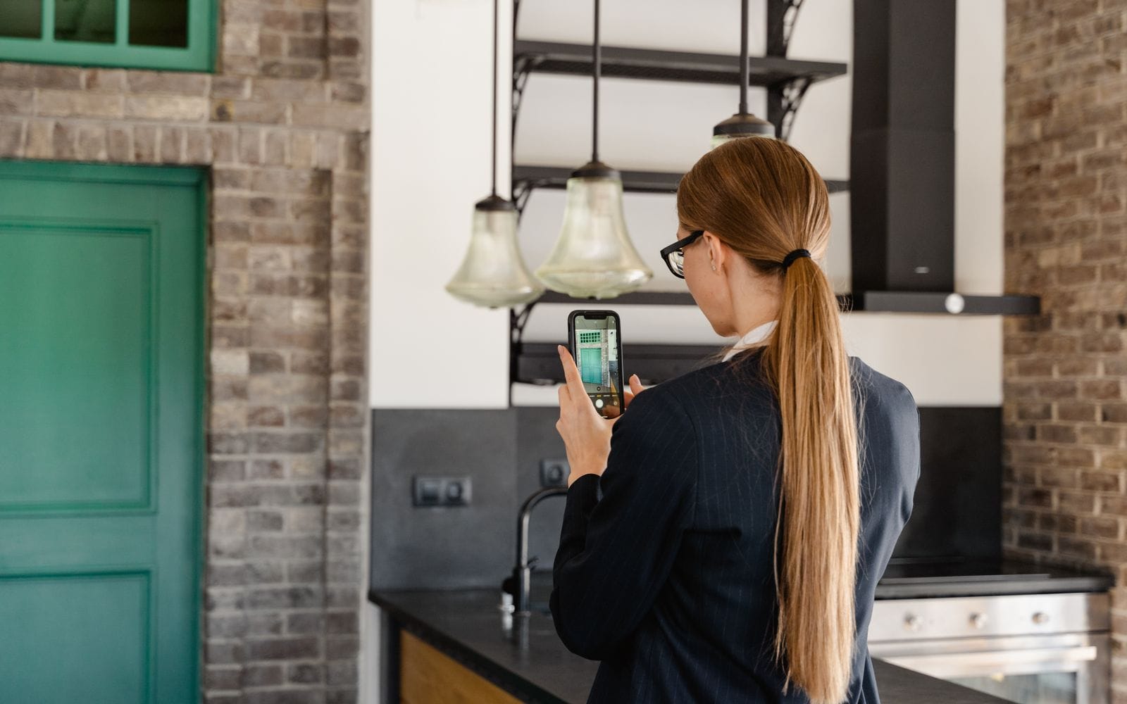 A woman in a dark blazer filming a modern kitchen with her smartphone