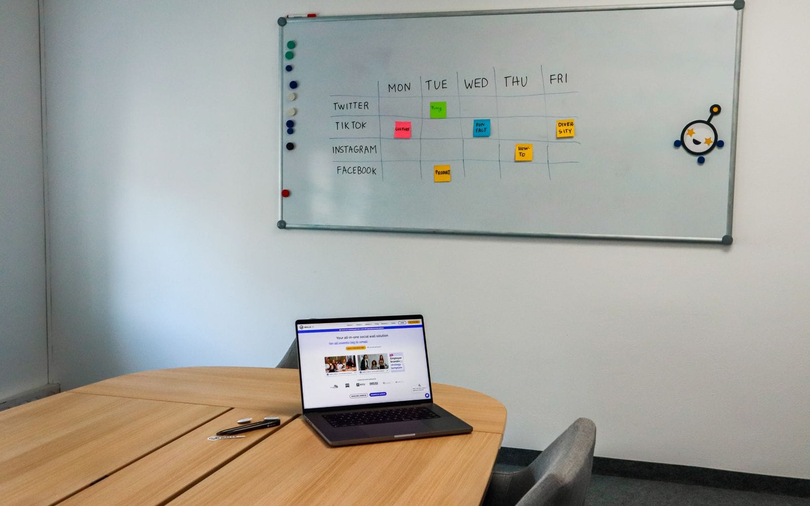 A whiteboard with a weekly social media content calendar above a laptop on a meeting table