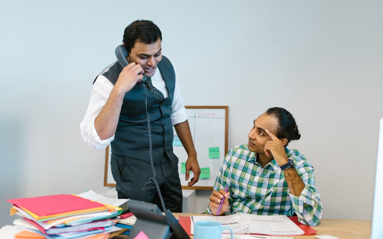 A man on a desk phone talking while a colleague listens in an office