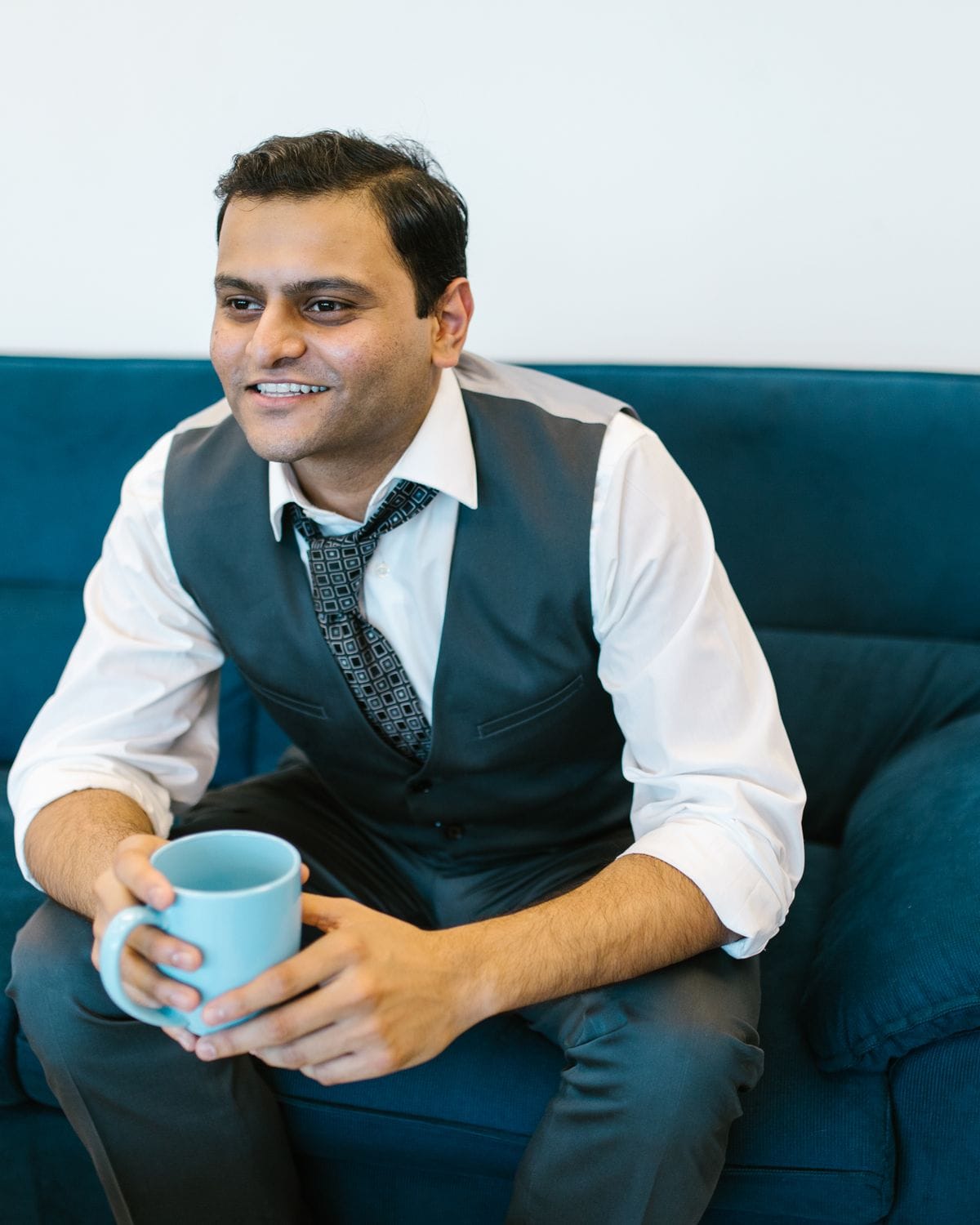 A smiling man in a waistcoat holding a mug during an informal office conversation