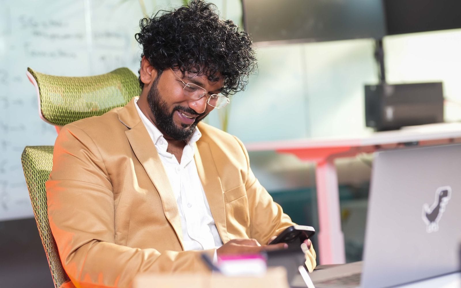 A man in a blazer smiling at his phone at a desk with a laptop
