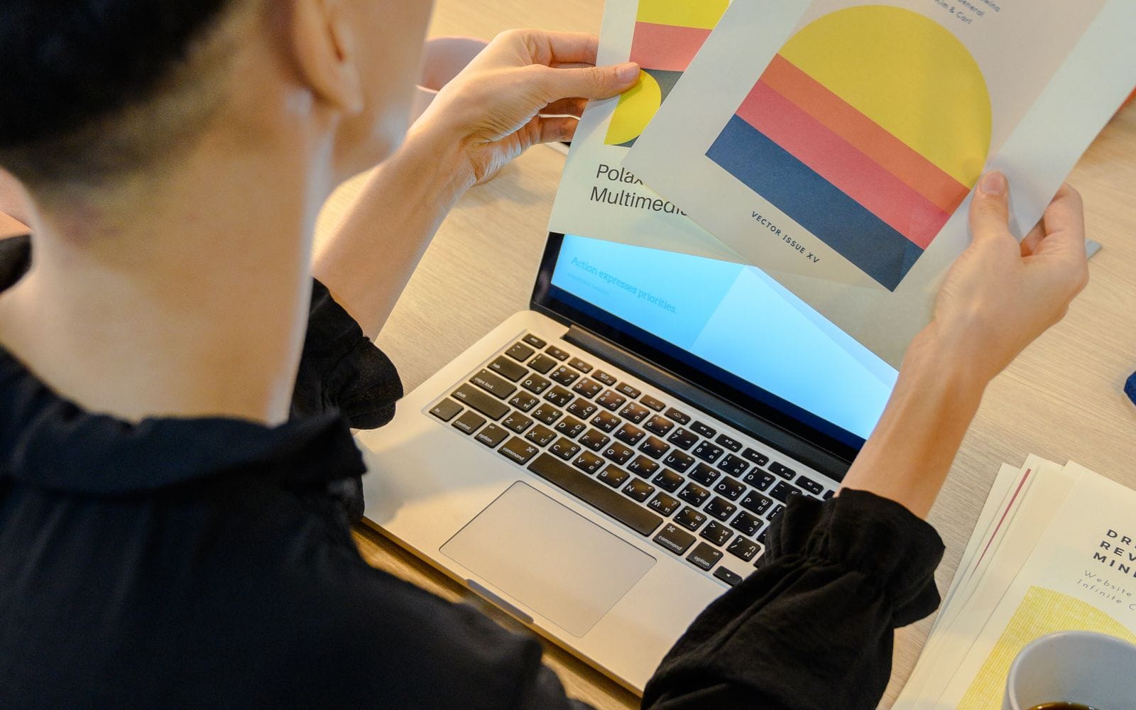 A designer holding up colourful printed brochure pages in front of a laptop
