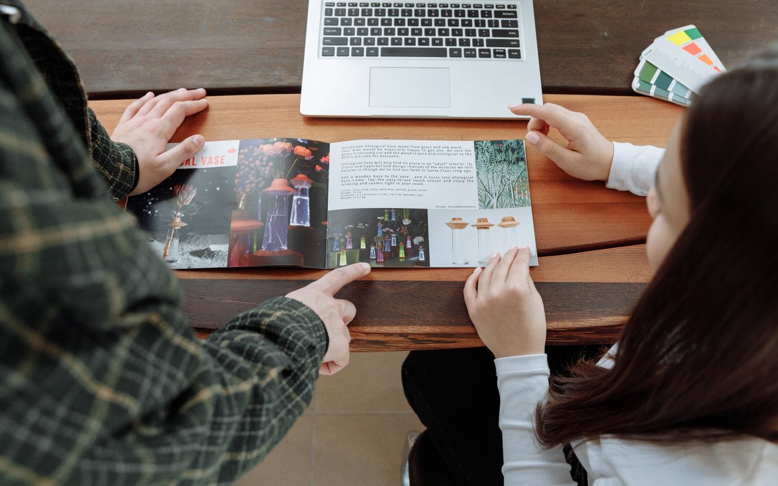 Two people pointing at pages of an open printed brochure on a wooden table