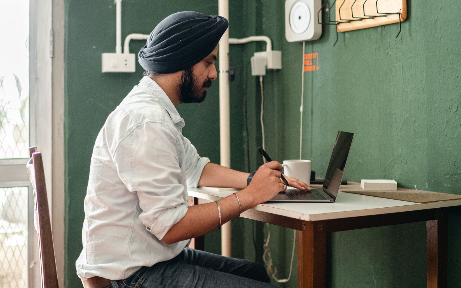 A man in a turban working on a laptop at a table by a window