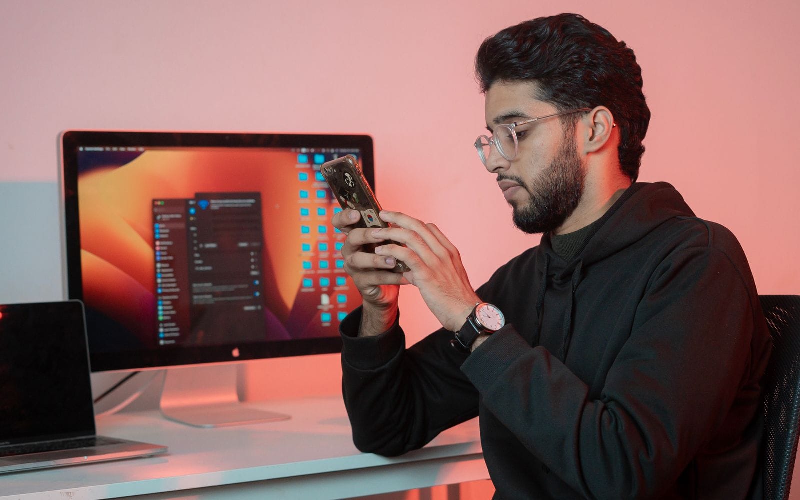 A man using his phone at a desk in front of a desktop monitor