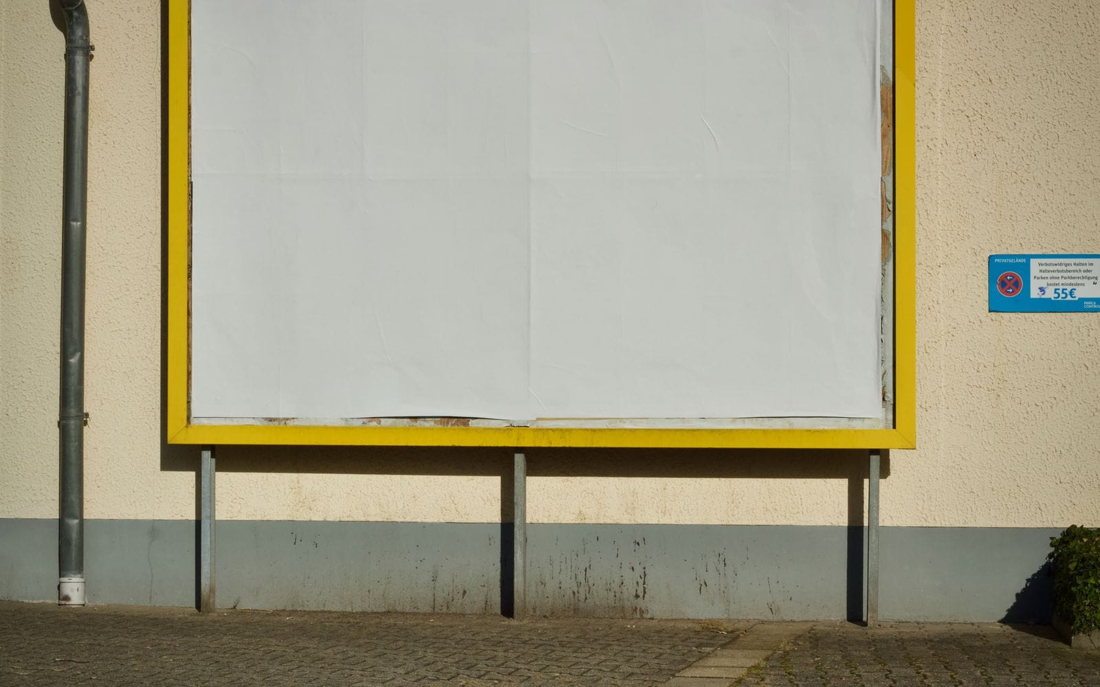 An empty poster board with a yellow frame fixed to a wall beside a pavement