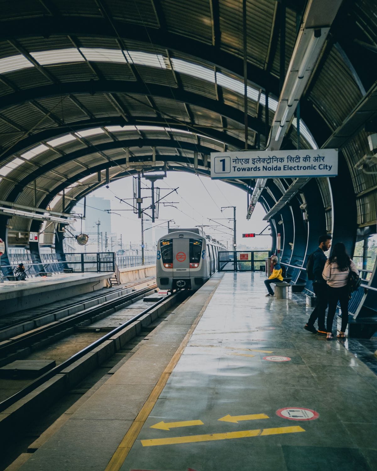 Passengers on an elevated metro platform as a train arrives, with a sign towards Noida Electronic City
